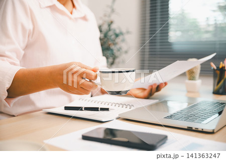Close-up office worker holding coffee and reviewing paper beside laptop, notebook, and phone on bright desk, showing business planning, finance report, remote work, productivity, 141363247