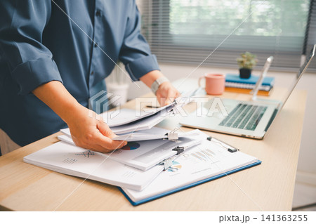 Hands organizing financial documents in a binder beside laptop and coffee on a bright office desk, showing accounting workflow, business report review, paperwork management, 141363255