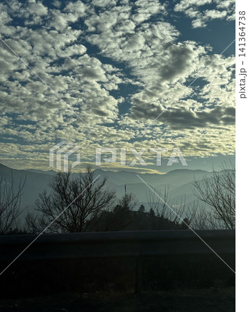 Mountain layers and cloudy sky viewed from roadside with trees in foreground. Mountain layers and cloudy sky viewed from roadside with trees in foreground. 141364738