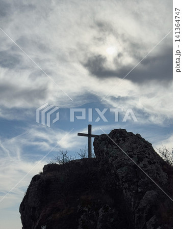 Cross on rocky peak beneath cloudy sky with bright sunlight diffused by clouds. 141364747