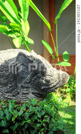 Carved stone guardian statue head framed by lush tropical leaves in Balinese garden with weathered relief, ornate patterns, sunlit textures, serene heritage atmosphere Carved stone guardian statue head framed by lush tropical leaves in Balinese garden with weathered relief, ornate patterns, sunlit textures, serene heritage atmosphere 141365187