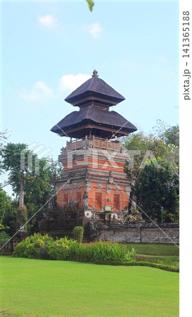Balinese temple tower with layered thatched roofs, red brick carvings, tropical trees, manicured lawn, and blue sky in Bali nature heritage garden setting, with balinese, temple, tower, thatched Balinese temple tower with layered thatched roofs, red brick carvings, tropical trees, manicured lawn, and blue sky in Bali nature heritage garden setting, with balinese, temple, tower, thatched 141365188