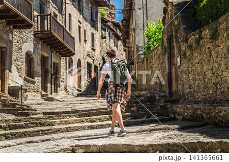 Young backpacker walking through historic stone street in Rupit village, Catalonia, Spain, exploring medieval architecture Young backpacker walking through historic stone street in Rupit village, Catalonia, Spain, exploring medieval architecture 141365661
