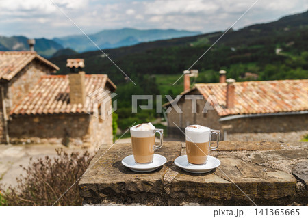 Two cappuccino cups on stone terrace overlooking peaceful mountain village Two cappuccino cups on stone terrace overlooking peaceful mountain village 141365665