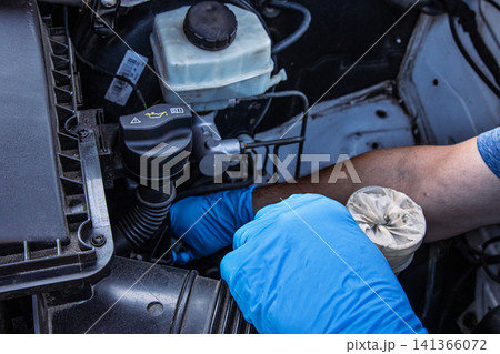 Auto mechanic wearing blue gloves holding a dirty car coolant reservoir filter, inspecting engine parts for maintenance and repair near an open vehicle hood. Fixing vehicle cooling system. Auto mechanic wearing blue gloves holding a dirty car coolant reservoir filter, inspecting engine parts for maintenance and repair near an open vehicle hood. Fixing vehicle cooling system. 141366072