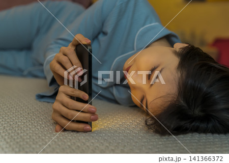 夜のベッドでスマホを見ながら横になる若い女性 夜のベッドでスマホを見ながら横になる若い女性 141366372