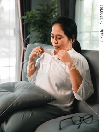 Middle aged Asian woman feeling hot and uncomfortable on sofa at home, pulling shirt collar with glasses nearby, concept of menopause hot flash and health discomfort. Middle aged Asian woman feeling hot and uncomfortable on sofa at home, pulling shirt collar with glasses nearby, concept of menopause hot flash and health discomfort. 141366594
