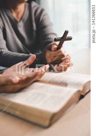Person holding a wooden cross above an open Bible with another open hand in prayer, Christian faith, blessing, worship, surrender, and hope concept. 141366651