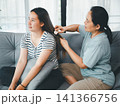 Asian mother gently brushing adult daughter hair on sofa at home, concept of family care, haircare, personal hygiene, bonding, caregiving, and daily lifestyle. 141366756