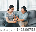 Asian mother and adult daughter checking smartwatch health tracker on sofa at home, concept of wearable technology, family care, digital healthcare, wellness, and support. 141366758