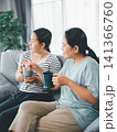 Asian mother and adult daughter sitting on sofa with cups at home, having conversation and pointing outside, concept of family communication, support, care, and trust. 141366760