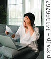 Middle aged Asian woman reading document beside laptop at home, touching forehead with headache and stress from paperwork, financial problem, online work, or health concern. 141366762