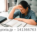 Asian woman sitting at table with notebook and glasses, touching forehead from headache, concept of stress, study, paperwork, work pressure, eye strain, and fatigue. 141366763