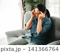 Middle aged Asian woman touching temples with eyes closed on sofa at home, concept of migraine, headache, stress, anxiety, burnout, fatigue, and health problem. 141366764