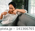 Asian woman feeling overheated and uncomfortable while sitting on sofa at home, pulling white shirt collar, concept of menopause, hot flash, sweating, and body heat. 141366765