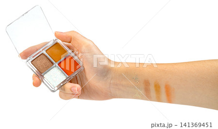 Hand holding a small four-color eyeshadow palette with orange, brown, and glitter shades. Makeup swatches applied to the forearm. Professional beauty product photography isolated. Hand holding a small four-color eyeshadow palette with orange, brown, and glitter shades. Makeup swatches applied to the forearm. Professional beauty product photography isolated. 141369451