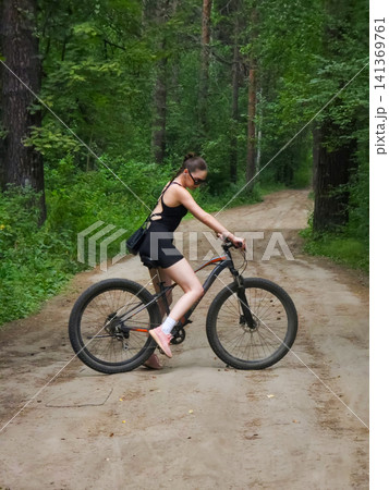 Caucasian woman riding a mountain bike on a dirt trail in a green forest. Active leisure lifestyle, outdoor exercise, and summer countryside travel. Sporty girl cycling in nature. Caucasian woman riding a mountain bike on a dirt trail in a green forest. Active leisure lifestyle, outdoor exercise, and summer countryside travel. Sporty girl cycling in nature. 141369761