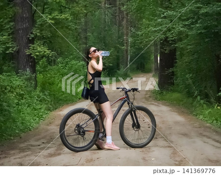Woman drinking water from bottle while sitting on bicycle in summer forest. Cyclist resting on park road during ride. Healthy lifestyle, outdoor sport activity and recreation concept. Woman drinking water from bottle while sitting on bicycle in summer forest. Cyclist resting on park road during ride. Healthy lifestyle, outdoor sport activity and recreation concept. 141369776