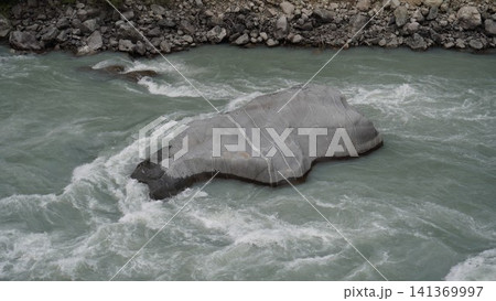 Large gray boulder partially submerged in rushing turquoise-white water and foam, geology concept. 141369997
