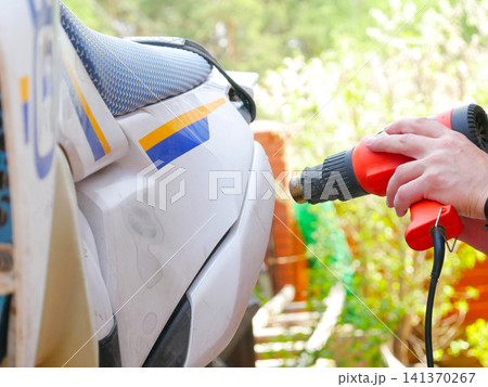 Man using heat gun to repair plastic motorcycle bodywork. Mechanic warming up and reshaping scratched fairing for vehicle restoration. Automotive maintenance and DIY garage repair service. 141370267