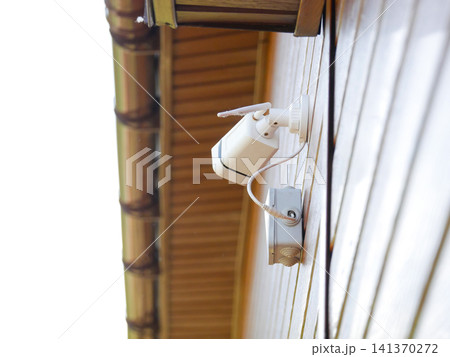 Security surveillance camera mounted on a wooden building wall. Outdoor CCTV system for home protection and property safety. Electronic device for monitoring activity and crime prevention. Security surveillance camera mounted on a wooden building wall. Outdoor CCTV system for home protection and property safety. Electronic device for monitoring activity and crime prevention. 141370272