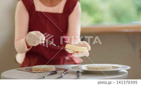 Young Asian woman spreading cream filling on cake layer with knife during home baking in bright kitchen 141370395