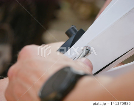 Man using screwdriver to tighten screw on white plastic frame. Professional handyman assembling furniture or repairing household equipment. DIY home improvement and construction work concept. 141370986