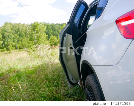 White car parked on grassy meadow edge. Driver door open in nature landscape. Travel concept, freedom of road trip and outdoor adventure. Summer automobile vehicle excursion lifestyle scene. White car parked on grassy meadow edge. Driver door open in nature landscape. Travel concept, freedom of road trip and outdoor adventure. Summer automobile vehicle excursion lifestyle scene. 141371598