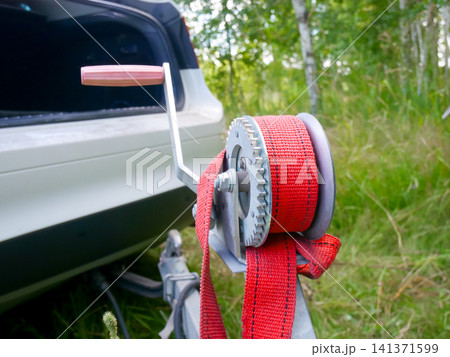 Manual boat trailer winch with red strap hooked to vehicle bumper used for transportation. Equipment for secure hauling of watercraft. Practical tool for outdoor and recreation activity. Manual boat trailer winch with red strap hooked to vehicle bumper used for transportation. Equipment for secure hauling of watercraft. Practical tool for outdoor and recreation activity. 141371599