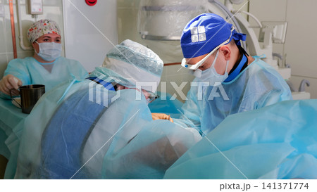 Surgical team in sterile gowns and masks performing medical procedure in operating room with blue drapes, clinical equipment, focused lighting, and careful teamwork during patient care Surgical team in sterile gowns and masks performing medical procedure in operating room with blue drapes, clinical equipment, focused lighting, and careful teamwork during patient care 141371774