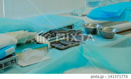 Sterile surgical instruments arranged on blue operating room drape with stainless steel bowls, metal trays, forceps, scissors, clamps, folded towels, and gauze prepared for clinical procedure 141371778