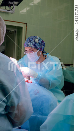Medical team works in sterile operating room as masked surgeon with protective eyewear handles surgical instruments over draped patient, illuminated by focused lights in clean hospital environment Medical team works in sterile operating room as masked surgeon with protective eyewear handles surgical instruments over draped patient, illuminated by focused lights in clean hospital environment 141371788