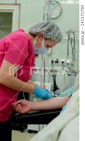 Masked nurse in scrubs preparing intravenous catheter for patient on treatment bed inside clean clinical room with infusion tubing, monitor, gloves, and bedside medical equipment Masked nurse in scrubs preparing intravenous catheter for patient on treatment bed inside clean clinical room with infusion tubing, monitor, gloves, and bedside medical equipment 141371795
