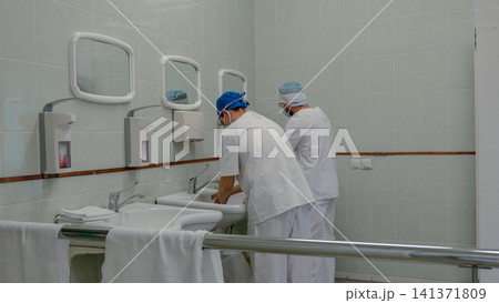 Medical staff washing hands in tiled hospital scrub room before surgery, wearing masks, caps, and white uniforms beside stainless steel sinks, mirrors, soap dispensers, and bright clinical lighting Medical staff washing hands in tiled hospital scrub room before surgery, wearing masks, caps, and white uniforms beside stainless steel sinks, mirrors, soap dispensers, and bright clinical lighting 141371809
