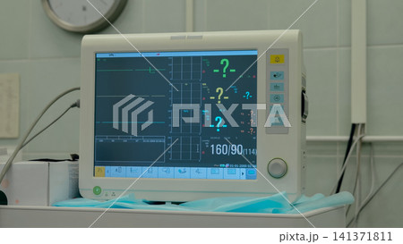Bedside patient monitor showing flat cardiac waveform and numeric vital readings on wheeled trolley in quiet clinical room with blue surgical drape, tiled wall, soft fluorescent light, and medical 141371811