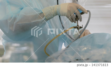 Gloved surgeon hands adjusting endoscopic instrument with clear tubing during minimally invasive procedure in sterile operating room with blue surgical drapes and bright clinical lighting Gloved surgeon hands adjusting endoscopic instrument with clear tubing during minimally invasive procedure in sterile operating room with blue surgical drapes and bright clinical lighting 141371813