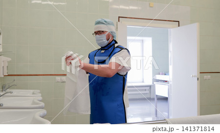 Masked medical worker wearing protective blue lead apron and cap stands in clean tiled hospital washroom while holding white coat near sinks with open doorway and soft clinical light Masked medical worker wearing protective blue lead apron and cap stands in clean tiled hospital washroom while holding white coat near sinks with open doorway and soft clinical light 141371814
