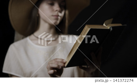 Young woman reading hardcover book under wide-brim hat, hands and pages in foreground with subtle page turn Young woman reading hardcover book under wide-brim hat, hands and pages in foreground with subtle page turn 141372274