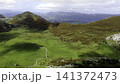 Sweeping aerial view of upland valley with winding footpath, bright pasture, narrow stream, rust-brown hills, shifting sunlight and calm rural landscape, with mountain, valley, pasture, meadow detail 141372473