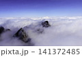 Jagged mountain peaks rising through drifting cloud deck beneath clear blue sky, dramatic alpine panorama with dark rock summits, mist, vast horizon, serene highland scale 141372485