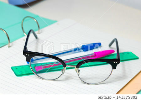 School supplies including glasses, two colorful pens, and a ruler laying on a lined open notebook, symbolizing readiness for learning, studying, and academic endeavors 141375802