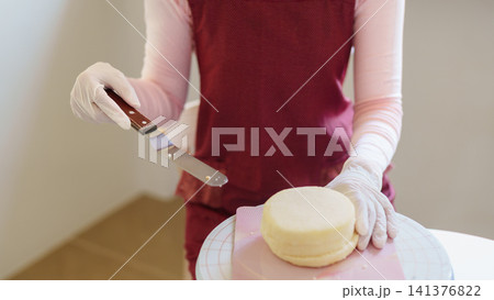 Asian baker wearing gloves spreading frosting on pink cake layer with palette knife on turntable Asian baker wearing gloves spreading frosting on pink cake layer with palette knife on turntable 141376822