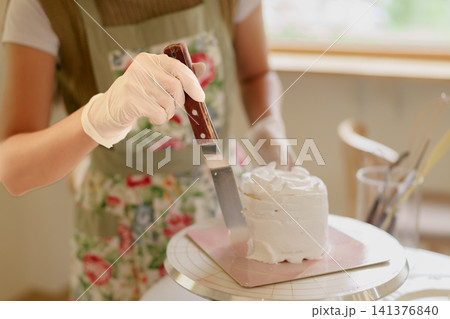 Baker smoothing white buttercream frosting on layered cake with metal spatula in bright home kitchen Baker smoothing white buttercream frosting on layered cake with metal spatula in bright home kitchen 141376840