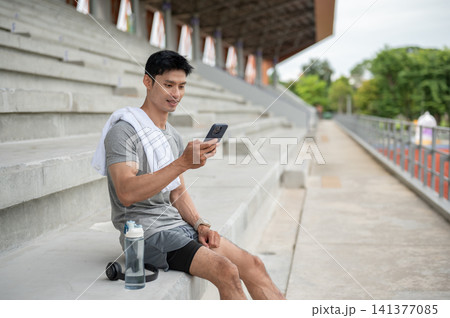 Asian athletic man sitting on seating stairs and using smartphone while resting in sports stadium. 141377085