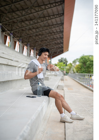 Smiling Asian man checking time on watch on grandstand steps in sports stadium. 141377090