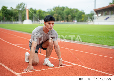 Asian male sprinter crouching in low start position on running track line in sports stadium. Asian male sprinter crouching in low start position on running track line in sports stadium. 141377099