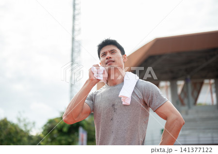 Tired Asian athlete wiping sweat with towel while standing in sports stadium. Tired Asian athlete wiping sweat with towel while standing in sports stadium. 141377122
