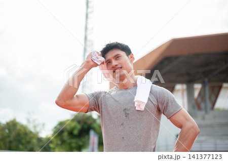 Asian man resting and wiping sweat with towel after running in sports stadium. Asian man resting and wiping sweat with towel after running in sports stadium. 141377123