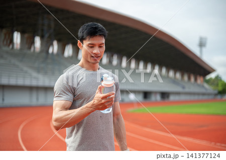 Asian athlete holding a water bottle while standing on running track in sports stadium. Asian athlete holding a water bottle while standing on running track in sports stadium. 141377124
