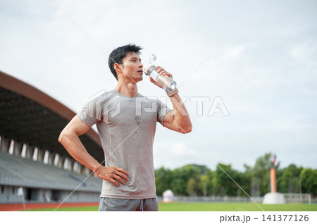 Thirsty Asian man drinking water from bottle while standing on track in sports stadium. Thirsty Asian man drinking water from bottle while standing on track in sports stadium. 141377126